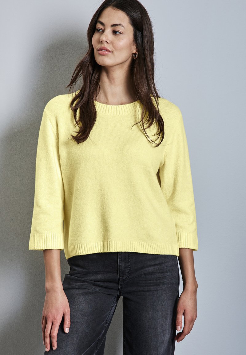 Frau mit langem dunklem Haar, die einen hellgelben Strickpullover mit drei Viertelärmeln und schwarze Jeans trägt, steht vor einer grauen Wand.
