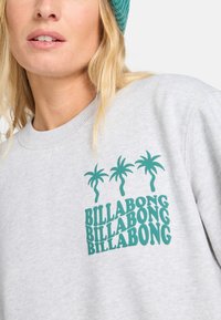 Grijze sweatshirt met groene palmboomafbeelding en "BILLABONG" tekst. De stof lijkt zacht met een ronde halslijn en geribde manchetten.