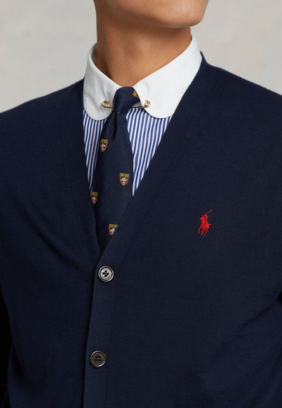 Cardigan in lana navy con scollo a V, cinque bottoni e logo polo rosso ricamato. Indossato sopra una camicia bianca con colletto e una cravatta a fantasia con scudi.