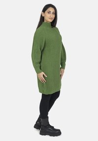 Abito maglione a coste verde con colletto alto, indossato sopra leggings neri e stivali neri con lacci spessi. Taglio semplice e rilassato.