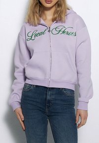 Liliowa bluza z kapturem na zamek z miękkiego materiału, z napisem "Local Heroes" w zielonym brokatowym foncie na klatce piersiowej, z przednimi kieszeniami i ściągaczami przy mankietach.