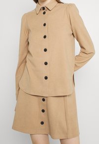 Abito beige con colletto, maniche lunghe e bottoni neri davanti. Tessuto morbido, design della gonna leggermente svasato.