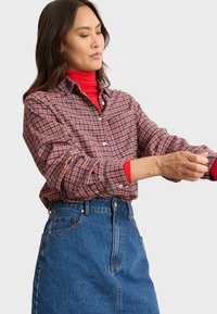 Chemise à carreaux rouge et marine à manches longues, portée par-dessus un col roulé rouge, accompagnée d'une jupe en denim taille haute.