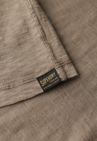 Tan tekstureret stof med synlige stikninger og et sort mærke med "Superdry" i guld. Materialet virker let og blødt.