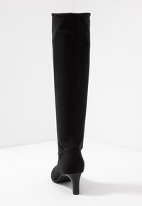 Bottes noires montantes au-dessus du genou en tissu extensible, présentant un talon effilé et une surface lisse et épurée sans quincaillerie ni accents visibles.