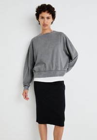 Sweatshirt cinza oversized com punhos canelados, usado sobre uma blusa branca, acompanhado de uma saia preta até ao joelho. Textura de tecido suave.