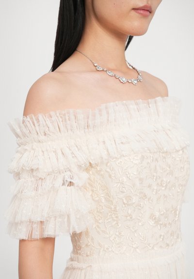 Sukienka z opuszczanymi ramionami w kolorze kremowym, z wielowarstwowymi falbanami na rękawach, haftowanymi kwiatowymi detalami oraz delikatnym srebrnym naszyjnikiem.