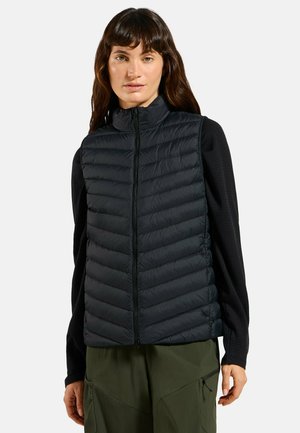 Frau trägt eine schwarze gesteppte Weste über einem schwarzen Langarmshirt und olivgrünen Hosen, steht vor einem einfachen hellen Hintergrund.