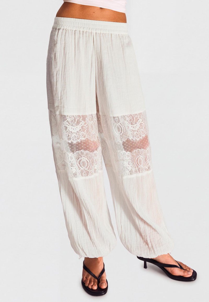 Pantalon blanc ample avec taille élastique et panneaux en dentelle aux genoux, porté avec des sandales à talons noires sur fond clair.