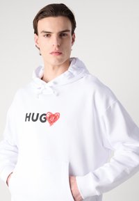 Hoodie bianco in cotone con tasca frontale, cappuccio con cordino e stampa "HUG" con un cuore rosso. Tessuto morbido, vestibilità comoda.