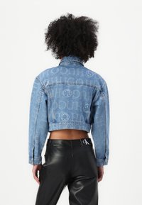 Veste en jean courte de couleur bleu clair avec un motif de logo répété. Dotée de poignets à boutons et d'un col classique, associée à un pantalon en cuir noir.