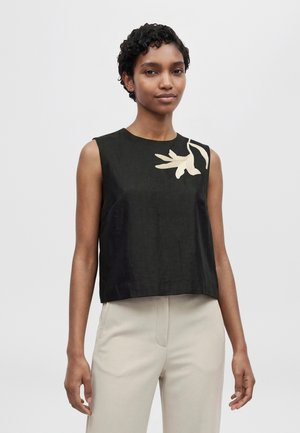 Un mannequin aux cheveux courts et bouclés porte un haut noir sans manches à motif floral crème et un pantalon clair sur un fond uni.