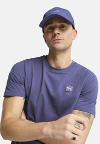 Camiseta de algodón morada con manga corta, que presenta un pequeño parche de logo blanco. Lleva una gorra de béisbol morada a juego.