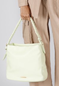 Světle zelená kožená kabelka s pleteným ramenním popruhem, měkkou texturou a hladkým povrchem. Na spodní části se nachází zlatý branding.