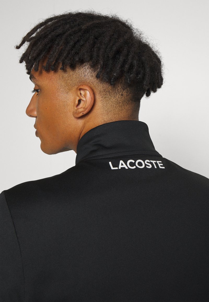 Zwarte poloshirt met een hoge kraag, voorzien van een witte "LACOSTE" logo aan de achterkant. De stof lijkt glad en lichtgewicht te zijn.