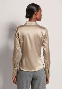 Beige satijnen overhemd met lange mouwen, een kraag en een getailleerde pasvorm, en met knoopmanchetten. De achtergrond toont een muur in een neutrale kleur.