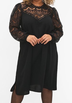 Femme portant une robe noire avec des manches longues en dentelle transparente et un empiècement en dentelle, debout les mains doucement jointes devant elle.