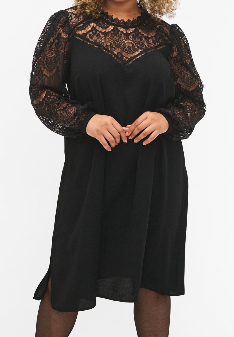 Femme portant une robe noire avec des manches longues en dentelle transparente et un empiècement en dentelle, debout les mains doucement jointes devant elle.
