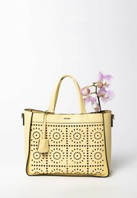 Gelbe Leder-Handtasche mit durchbrochenem geometrischem Muster, schwarzen Akzenten, zwei Griffen und einem abnehmbaren Anhänger. Ein Stängel lila Orchideen befindet sich darin.
