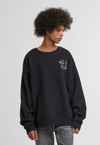Sweatshirt noir avec un col rond, des manches longues et un logo brodé sur la poitrine ; il présente une coupe décontractée et surdimensionnée.