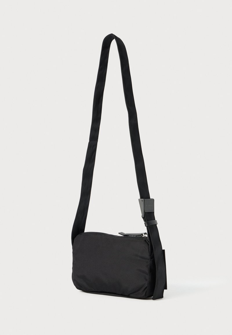 Sac à bandoulière rectangulaire noir avec une longue sangle ajustable et une fermeture éclair, présenté sur un fond clair uni.