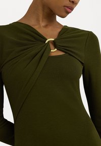 Olivgrön långärmad topp i ribbat tyg, med en vriden framsida och en gyllene cirkulär detalj vid halsen.