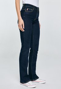 Osoba u tamnoplavim trapericama širokih nogavica i bijelim tenisicama stoji na jednostavnoj svijetloj pozadini.