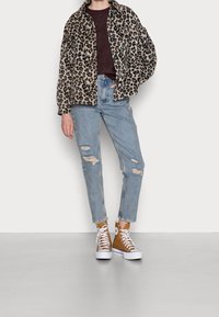 Veste en fausse fourrure motif léopard, pull en tricot bordeaux, jean bleu clair déchiré, et baskets montantes camel avec semelles blanches.