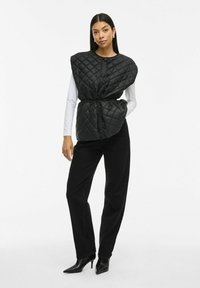 Svart quiltad väst med halvcirkelformade axlar, framknappar, justerbar dragsko i midjan, bärs över en vit skjorta, kombinerad med svarta byxor och klackar.