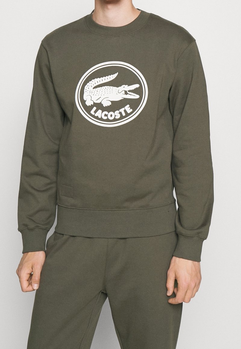 Sweatshirt vert olive à manches longues. Présente un logo crocodile Lacoste blanc dans un motif circulaire. Tissu doux avec poignets et ourlet côtelés.