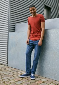 Rød bomull T-skjorte, blå denimjeans og marineblå sneakers. Modellen står mot en grå metallvegg, med en avslappet passform og enkelt design.