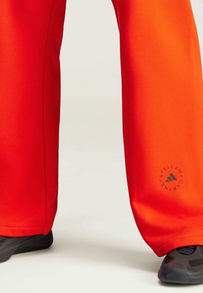 Pantaloni sportivi rossi con una vestibilità rilassata, caratterizzati da un logo grigio sulla parte inferiore della gamba e indossati con scarpe da ginnastica nere. Texture liscia e design moderno.