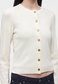 Femme portant un cardigan crème boutonné avec des boutons dorés et un jean foncé, montrée du cou à la taille.