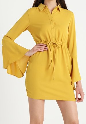 Femme portant une robe courte jaune moutarde à manches longues évasées, avec col, boutons sur le devant et taille à cordon.