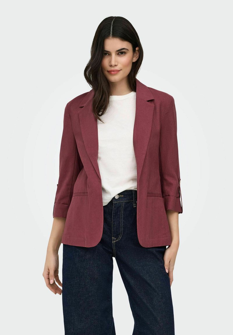 Femme aux longs cheveux foncés portant un blazer bordeaux avec les manches retroussées sur un t-shirt blanc et un jean taille haute bleu foncé.