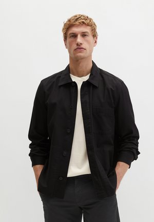 Młody mężczyzna o blond włosach, ubrany w czarną koszulę zapinaną na guziki na białej koszulce, stojący z rękami w kieszeniach na jednolitym tle.