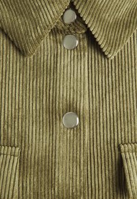 Giacca in corduroy verde oliva con costine larghe, lucidi bottoni rotondi argentati, colletto appuntito e tasche a toppa frontali.