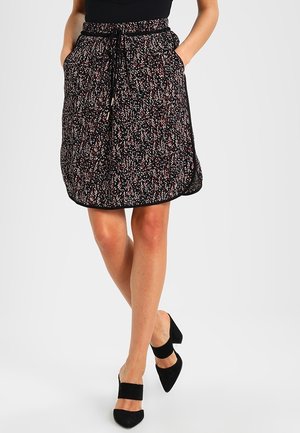 Femme portant une jupe noire à fleurs mi-longue avec des poches, un haut noir et des chaussures à talons noires, debout avec les jambes croisées.