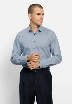 Mann trägt ein hellblau gemustertes, langärmliges Hemd mit Knöpfen und schwarze Hose, richtet seine linke Manschette und schaut nach rechts