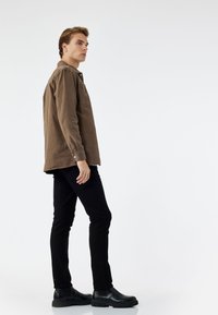 Braune Cordjacke mit Brusttasche, kombiniert mit schmalen schwarzen Jeans und schwarzen Stiefeletten vor einem schlichten weißen Hintergrund.