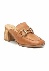 Mule de cuero marrón con punta redonda, detalle decorativo de hardware dorado y un tacón de bloque texturizado. La plantilla presenta el nombre de la marca 'Carmela.'