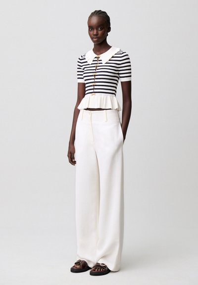 Mujer con una blusa de manga corta a rayas blancas y negras con bajo con volantes y pantalones blancos de pierna ancha, de pie frente a un fondo liso.