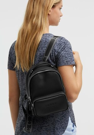 Woman with long blonde hair wears a black textured leather backpack over a short-sleeve patterned gray shirt, facing sideways on a white background.