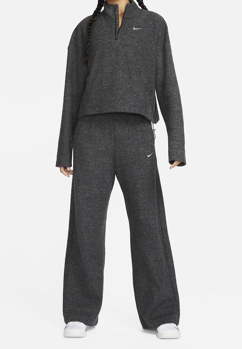 Personne portant un survêtement Nike gris foncé à manches longues et pantalon à jambes larges, assorti à des baskets blanches sur un fond uni.