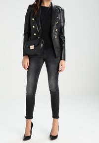 Donna che indossa un blazer di pelle nera con bottoni dorati, top nero, jeans skinny neri sbiaditi, tacchi neri e borsa a tracolla nera.
