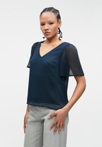 Ναυτικό μπλουζάκι με V-κοψη, κοντά διάφανα φουσκωτά μανίκια, ελαφρώς χαλαρή εφαρμογή και λεία υφή, σε συνδυασμό με ανοιχτό γκρι τζιν.