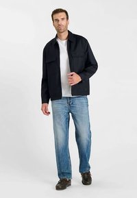 Marineblauw jack met twee borstzakken, gedragen over een wit T-shirt. Lichtgewassen jeans met rechte pijpen en bruine schoenen. Model staat tegen een effen achtergrond.