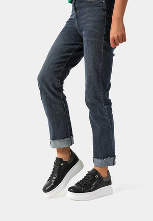 Sneaker in pelle nera con suola piatta bianca, dotati di occhielli dorati, lacci e un accento grigio sul tallone. Indossati con jeans scuri risvoltati.