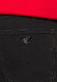 Czarne jeansy z denimu z naszytą kieszenią ozdobioną wypukłym, tłoczonym logo orła. Czerwony materiał widoczny nad paskiem.