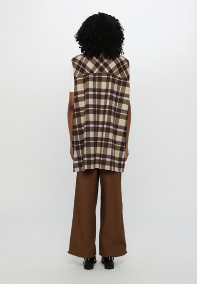 Top sin mangas a cuadros en tonos beige y marrón con un cuello puntiagudo, combinado con pantalones de pierna ancha marrones y zapatos negros, visto desde la espalda.
